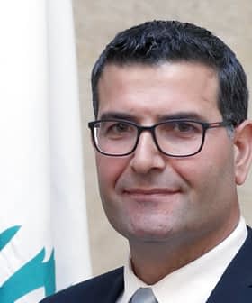 Man wearing glasses and a suit, standing in front of a Lebanese flag. - Olive Oil Times