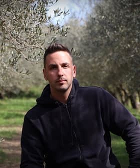 A man wearing a black hoodie sitting among olive trees in an olive grove. - Olive Oil Times