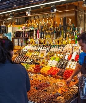 A market stall displaying various dried fruits, nuts, and bottles of olive oil with a vendor assisting a customer. - Olive Oil Times