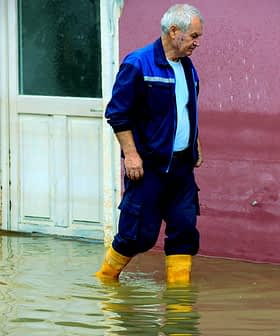 An older man wearing yellow boots walking through a flooded area near a building. - Olive Oil Times