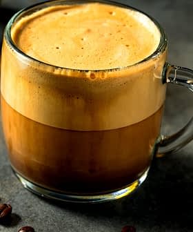 A glass mug filled with coffee featuring a distinct layer of cream on top, surrounded by coffee beans. - Olive Oil Times