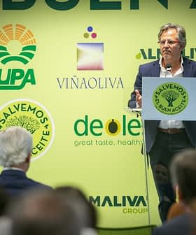 A speaker presenting at an olive oil industry event with a green backdrop featuring various logos. - Olive Oil Times