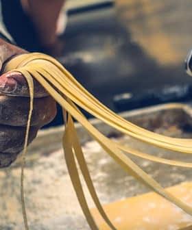 A hand holding freshly rolled pasta strands from a pasta machine in a kitchen setting. - Olive Oil Times