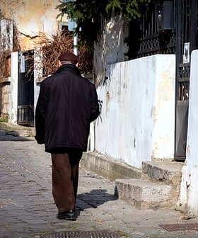An elderly man walking away on a cobblestone street lined with buildings and greenery. - Olive Oil Times