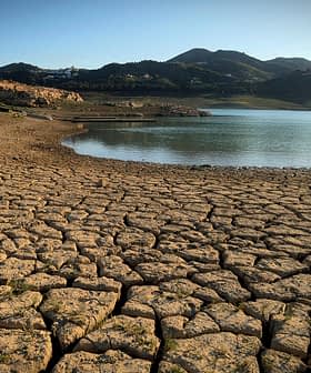 Dry, cracked earth surrounding a small body of water with mountains in the background. - Olive Oil Times