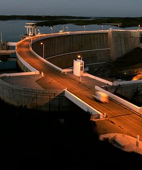 Concrete dam structure with a roadway and lights at dusk, reflecting in the water below. - Olive Oil Times