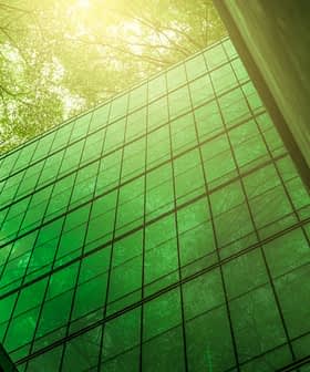 A modern glass building reflecting green light from surrounding trees in an urban setting. - Olive Oil Times