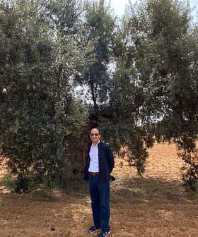 A man wearing sunglasses standing next to a large olive tree in a field. - Olive Oil Times
