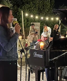 A woman singing into a microphone while seated at a piano during a performance at night. - Olive Oil Times