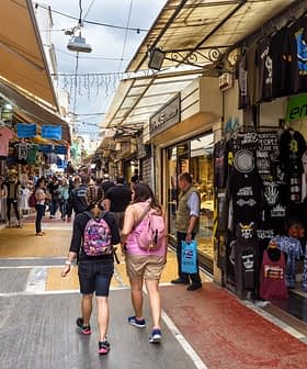 Two people walking through a market street lined with shops and stalls selling various items. - Olive Oil Times