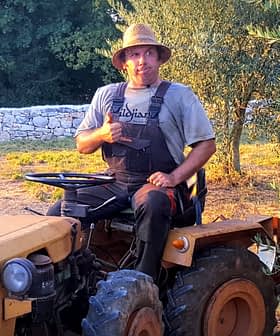A man sitting on a yellow tractor wearing a straw hat and overalls, giving a thumbs up. - Olive Oil Times