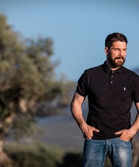 Man wearing a black polo shirt and jeans standing outdoors with olive trees in the background. - Olive Oil Times