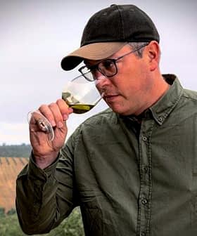 Man in a green shirt and cap tasting olive oil from a glass in a vineyard setting. - Olive Oil Times
