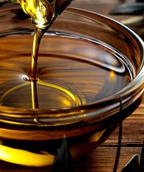 Golden olive oil being poured into a glass bowl placed on a wooden surface. - Olive Oil Times