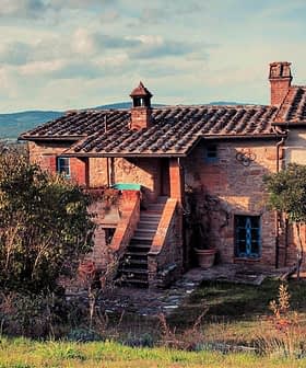 Two-story farmhouse with a tiled roof and blue window shutters surrounded by greenery. - Olive Oil Times