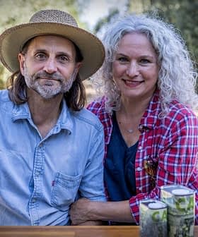 A man and woman sitting together outdoors, both smiling, with cans placed on a table in front of them. - Olive Oil Times