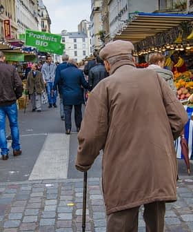 Elderly man with a cane walking through a market street lined with stalls and produce. - Olive Oil Times
