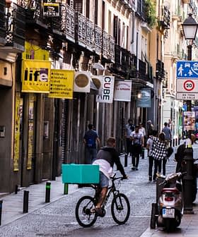 A street scene featuring a bicyclist with a teal delivery box and pedestrians walking in a narrow urban area. - Olive Oil Times