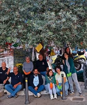 A group of people gathered under a large olive tree, some wearing gloves and smiling at the camera. - Olive Oil Times