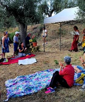 Group of people gathered outdoors under a tent for a performance, with some seated on blankets and others standing. - Olive Oil Times