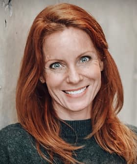 Woman with red hair smiling while leaning against a textured wall. - Olive Oil Times
