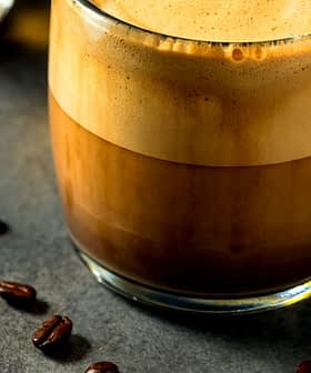 A glass mug filled with a layered coffee beverage, featuring a frothy top and dark liquid below. - Olive Oil Times