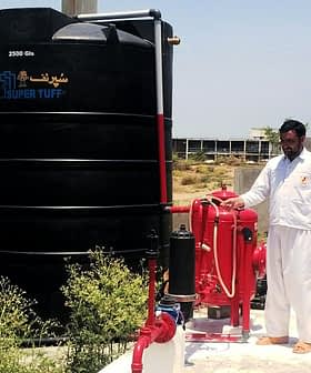 A large black water storage tank labeled 'SUPER TUFF' next to a red pump system and a man in white clothing. - Olive Oil Times
