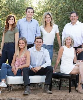 Seven individuals posing together outdoors in a natural setting with olive trees in the background. - Olive Oil Times