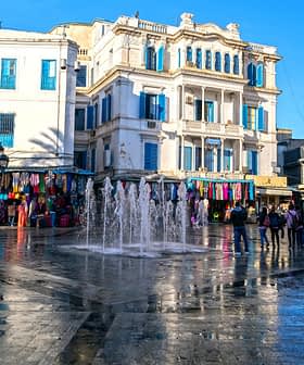 A fountain in a public square surrounded by buildings and people walking. - Olive Oil Times