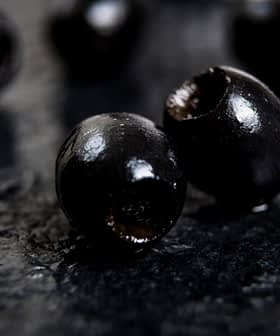 Close-up of black olives placed on a dark textured surface. - Olive Oil Times