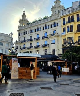 Wooden market stalls set up in an urban square with buildings in the background. - Olive Oil Times