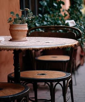 A round table with a potted plant and three chairs in a narrow outdoor setting. - Olive Oil Times