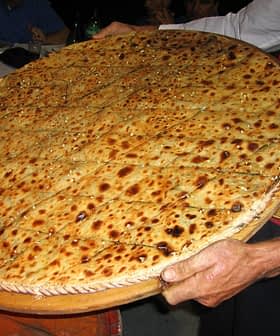 A person holding a large round flatbread topped with seeds and spices, presented on a wooden tray. - Olive Oil Times