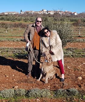 A couple standing in an olive grove with a dog, smiling at the camera. - Olive Oil Times