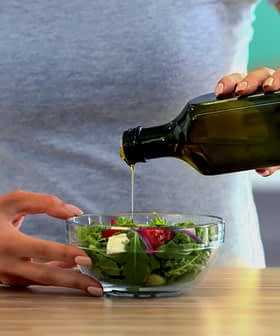Individual pouring olive oil from a dark glass bottle onto a fresh salad in a clear bowl. - Olive Oil Times