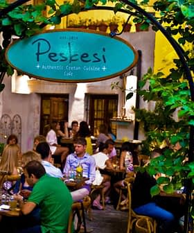 Entrance sign of Peskesi restaurant with greenery and patrons dining inside. - Olive Oil Times