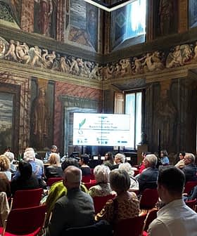 Audience seated in a historic hall during a conference presentation with decorative walls and a large screen. - Olive Oil Times