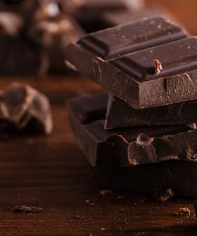 Several pieces of dark chocolate stacked on a wooden surface with additional chocolate fragments scattered around. - Olive Oil Times