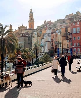 People walking along a promenade in a coastal town with colorful buildings and palm trees. - Olive Oil Times