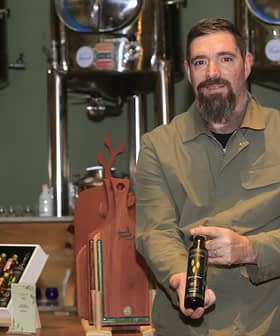 Man with a beard holding a bottle of olive oil in a production setting with equipment in the background. - Olive Oil Times