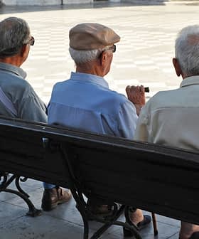 Three elderly men sitting on a park bench, viewed from behind, wearing hats and light-colored shirts. - Olive Oil Times