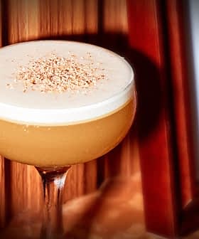 A cocktail in a coupe glass with a frothy top, garnished with grated nutmeg, is placed on a wooden surface next to a framed picture. The background is a wooden panel wall.