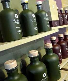 Verschiedene Olivenölflaschen aus grünem und braunem Glas mit Holzverschlüssen auf einem Regal angeordnet. – Olive Oil Times