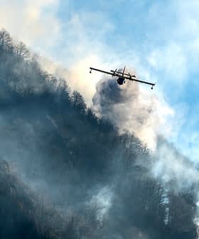 Uma aeronave liberando água para combater um incêndio florestal em uma região montanhosa com fumaça subindo do solo. - Olive Oil Times