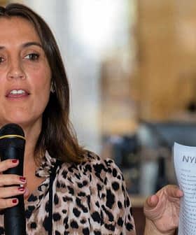 Frau hält ein Mikrofon und ein Blatt Papier, während sie bei einer Veranstaltung spricht. - Olive Oil Times