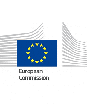 Logotipo de la Comisión Europea con fondo azul con estrellas amarillas y contornos de edificios estilizados. Olive Oil Times