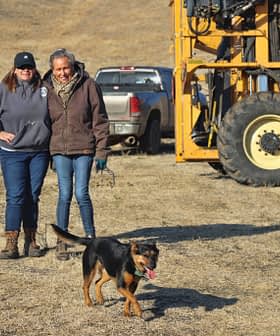 Deux femmes debout à côté d'une machine jaune avec un chien courant devant elles dans un champ ouvert. - Olive Oil Times