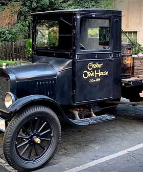 Ένα vintage μαύρο pickup truck με ξύλινα βαρέλια στο πίσω μέρος, παρκαρισμένο σε μια πλακόστρωτη επιφάνεια. - Olive Oil Times