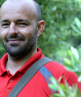 Un homme portant un polo rouge debout parmi les oliviers avec un léger sourire. - Olive Oil Times