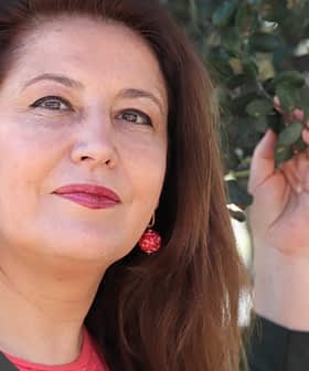 Femme aux longs cheveux bruns portant des boucles d'oreilles rouges, posant à l'extérieur avec une main sur une branche d'arbre. - Olive Oil Times